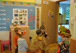 Piątka dzieci w jesiennych przebraniach stoi razem w sali przedszkolnej obok drzwi.